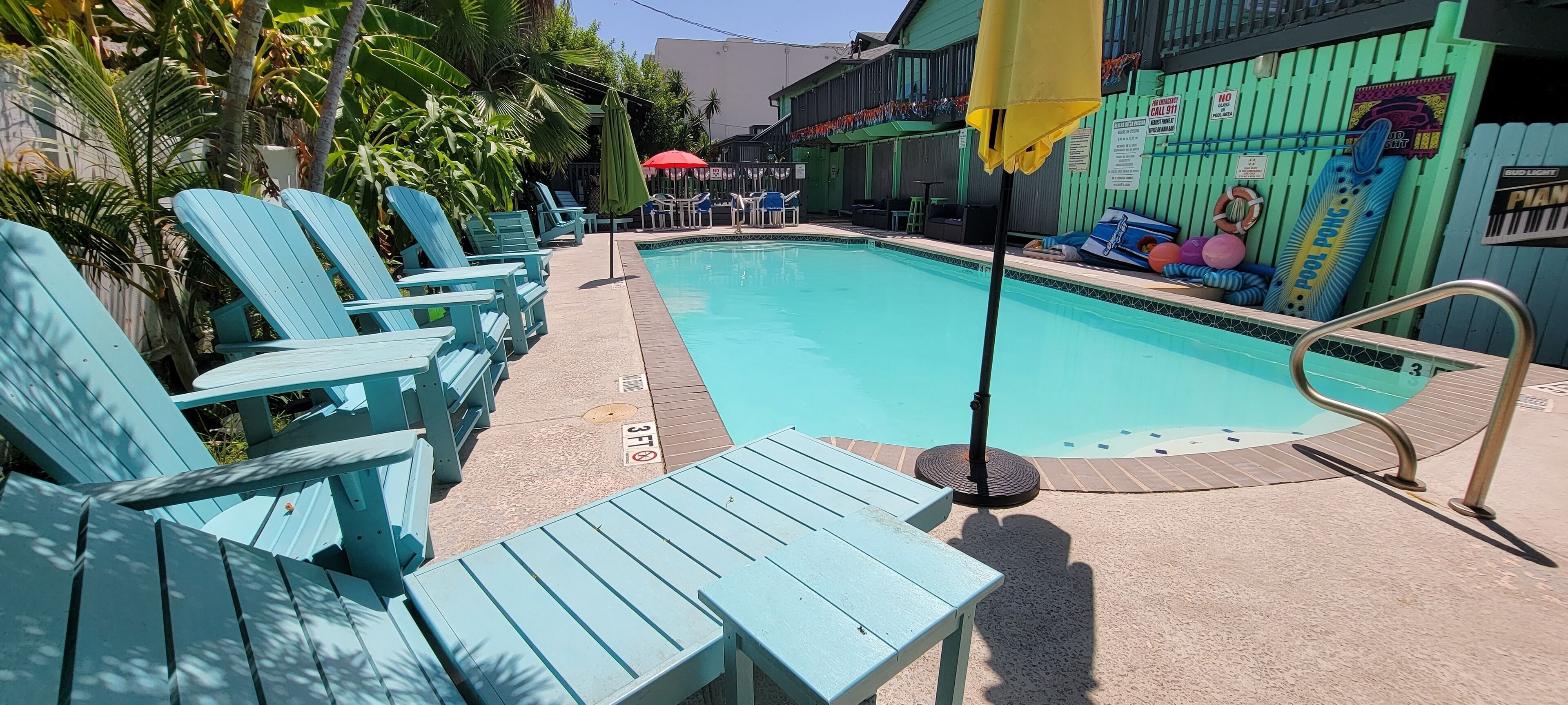 Zona de piscina con sillas azules y mesa de billar, rodeada por una valla verde y plantas tropicales.