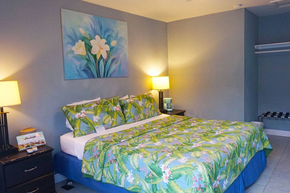 A hotel room with a bed, two bedside lamps, a painting, a small table with a coffee maker, and a chair.