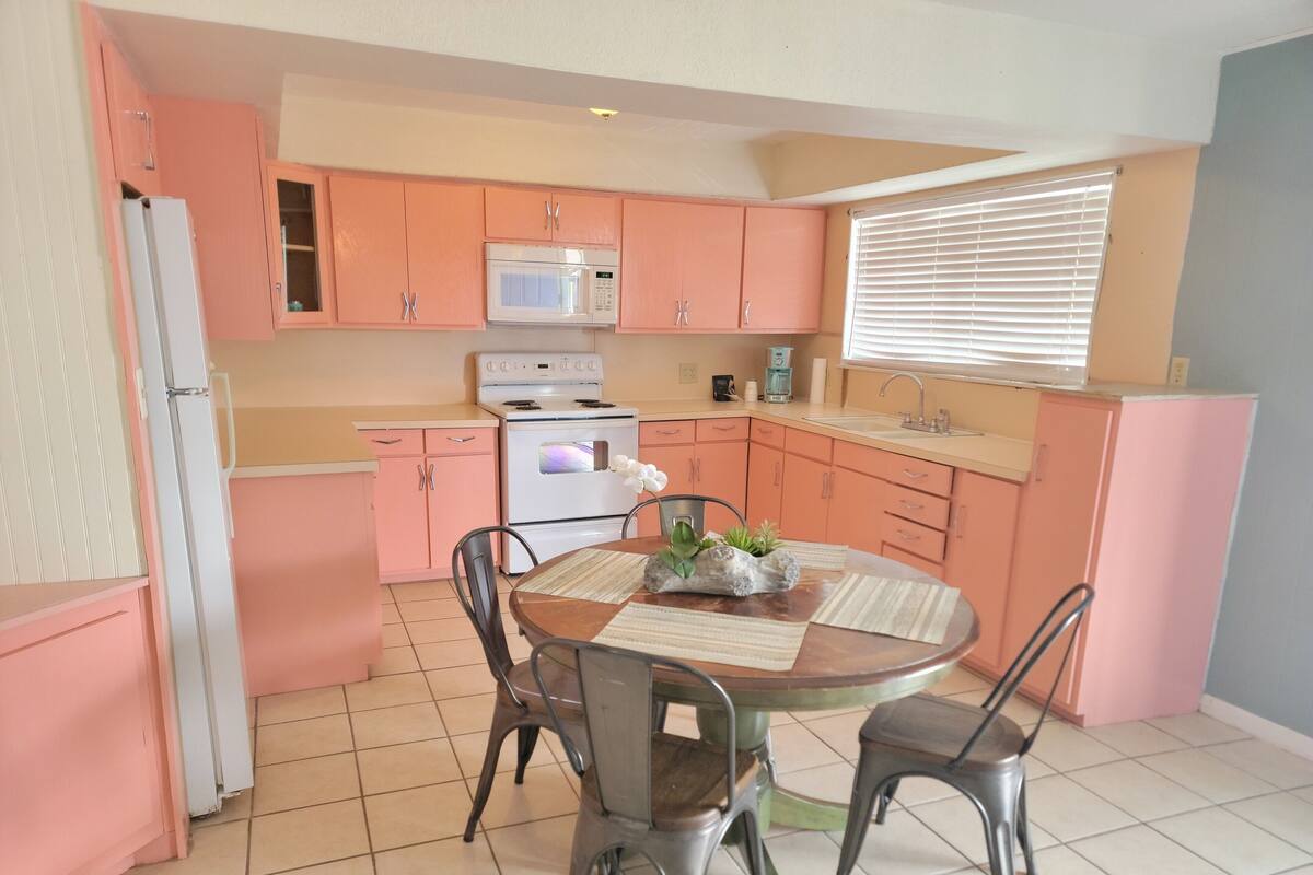 A kitchen with pink cabinets, a white refrigerator, a microwave, an oven, and a sink. There is a dining table with chairs and a vase with flowers.