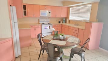 A kitchen with pink cabinets, a white refrigerator, a microwave, an oven, and a sink. There is a dining table with chairs and a vase with flowers.
