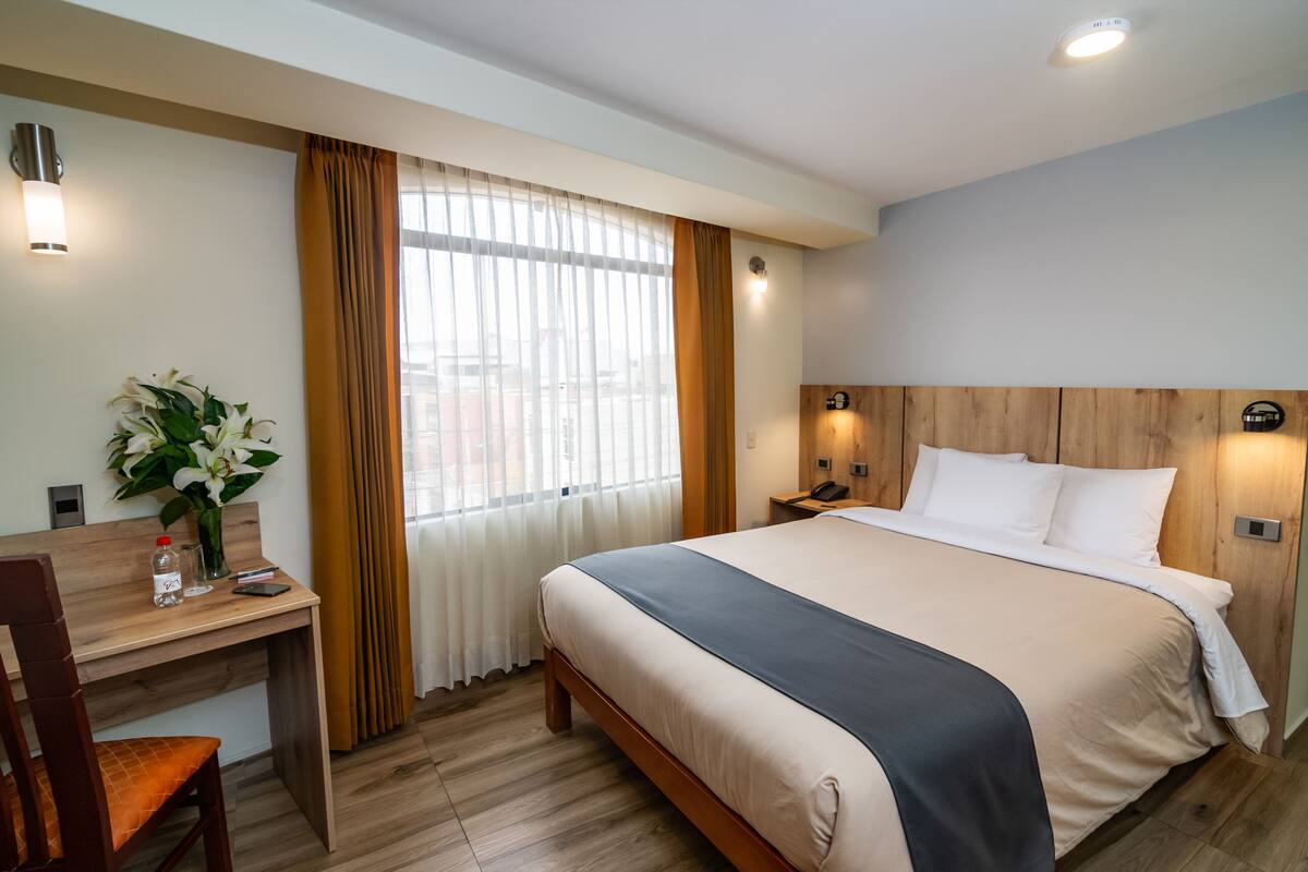 Ett hotellrum med en säng, ett skrivbord med en stol, en vas med blommor och ett fönster med gardiner.
