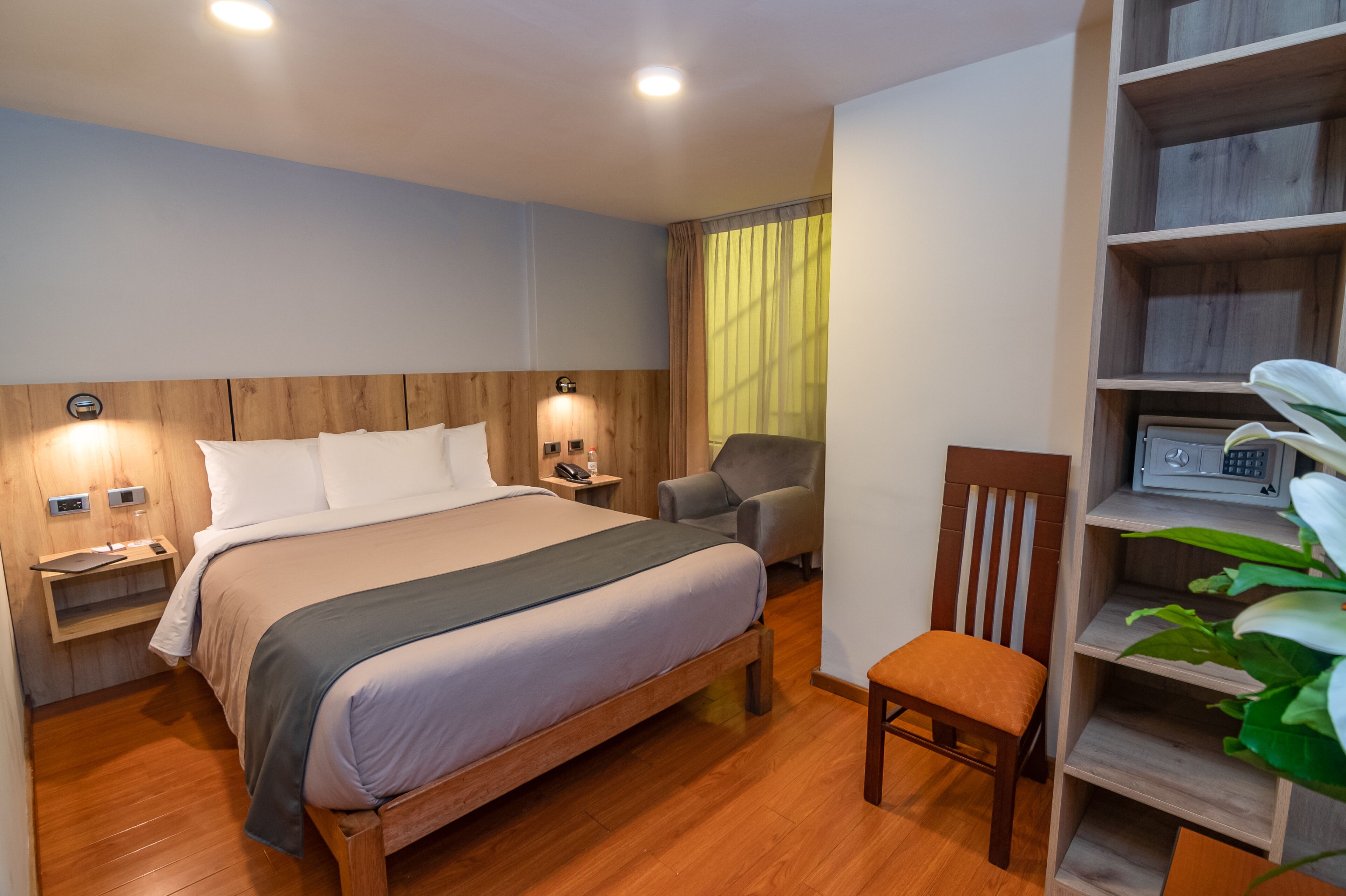 A hotel room with a wooden bed, a chair, a desk, and a shelf with a plant.