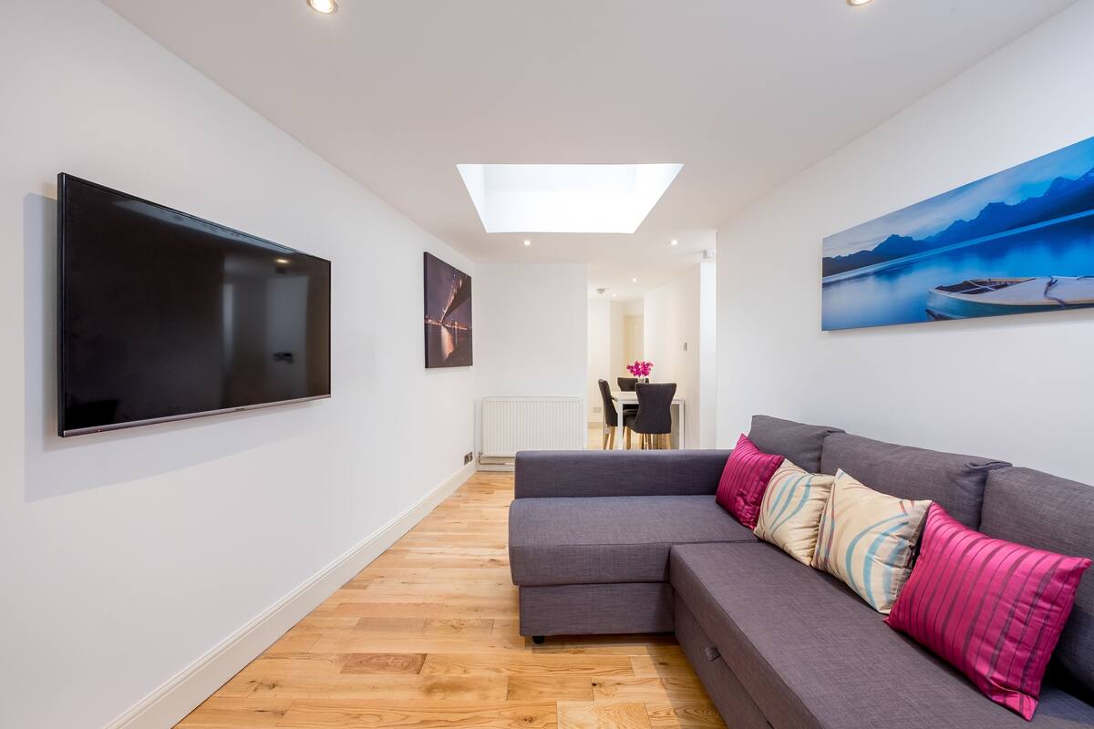 A modern living room with a flat-screen TV, a grey sofa with patterned cushions, wooden flooring, and a skylight.