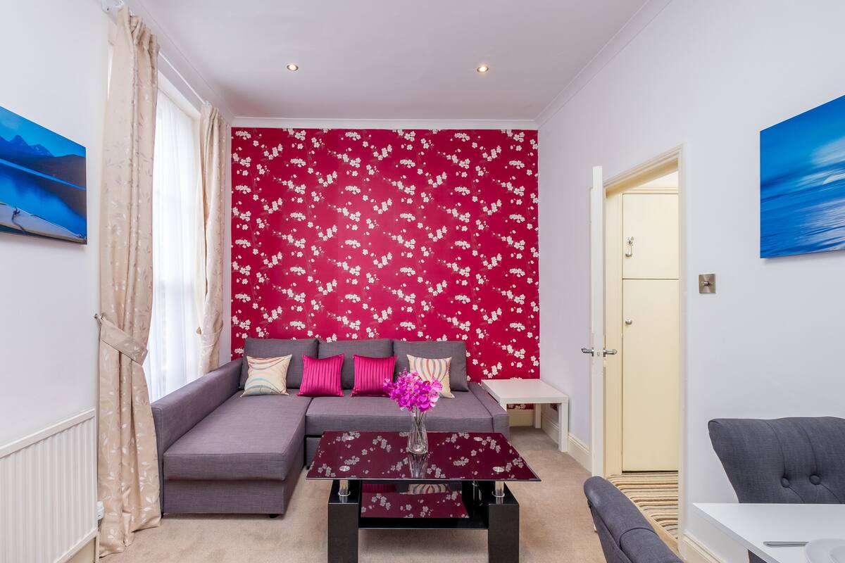 A modern living room with a grey sofa, a glass coffee table with a vase of flowers, and a red floral wallpaper.
