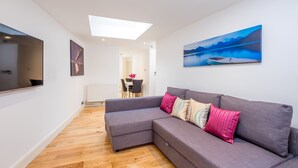 A modern living room with a grey sectional sofa, a flat-screen TV, a painting of a boat on water, and a skylight. - Marylebone Apartments (London)