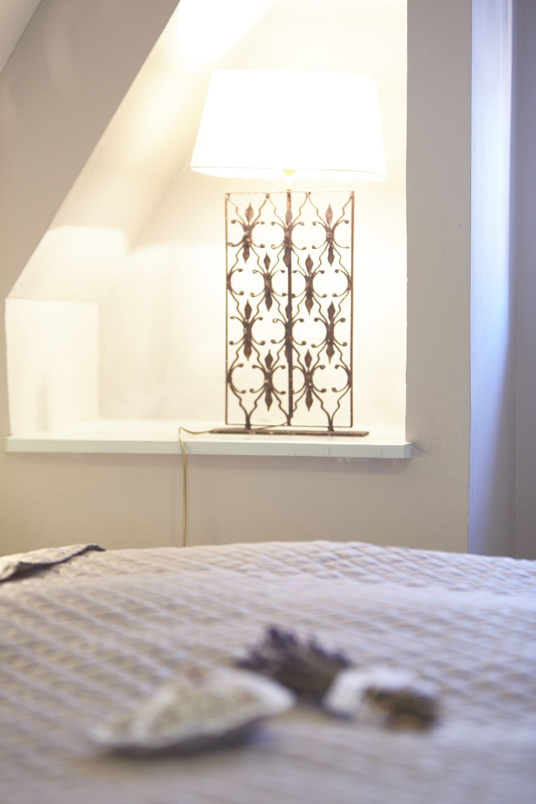 A bedroom with a bed, a window with a decorative metal frame, and a bedside table with a lamp.