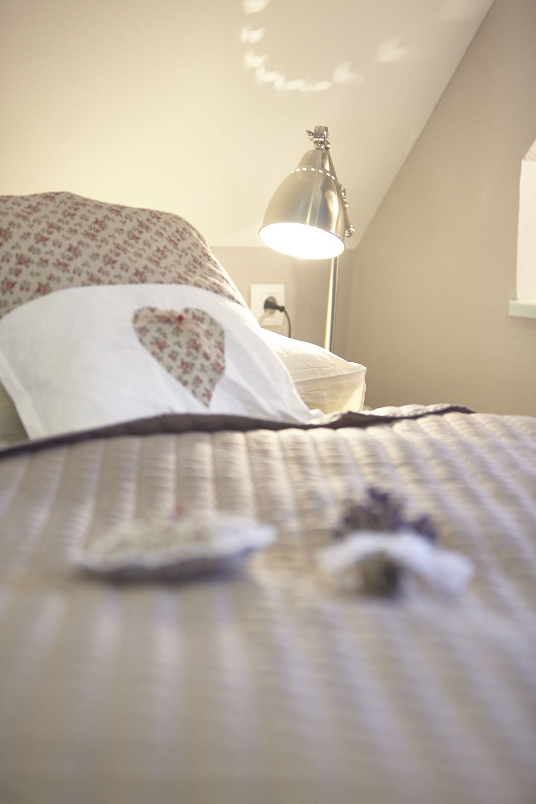 Un lit avec une couvre-lit à motifs et un oreiller en forme de cœur. Il y a une lampe sur une table de chevet et une fenêtre avec des rideaux.
