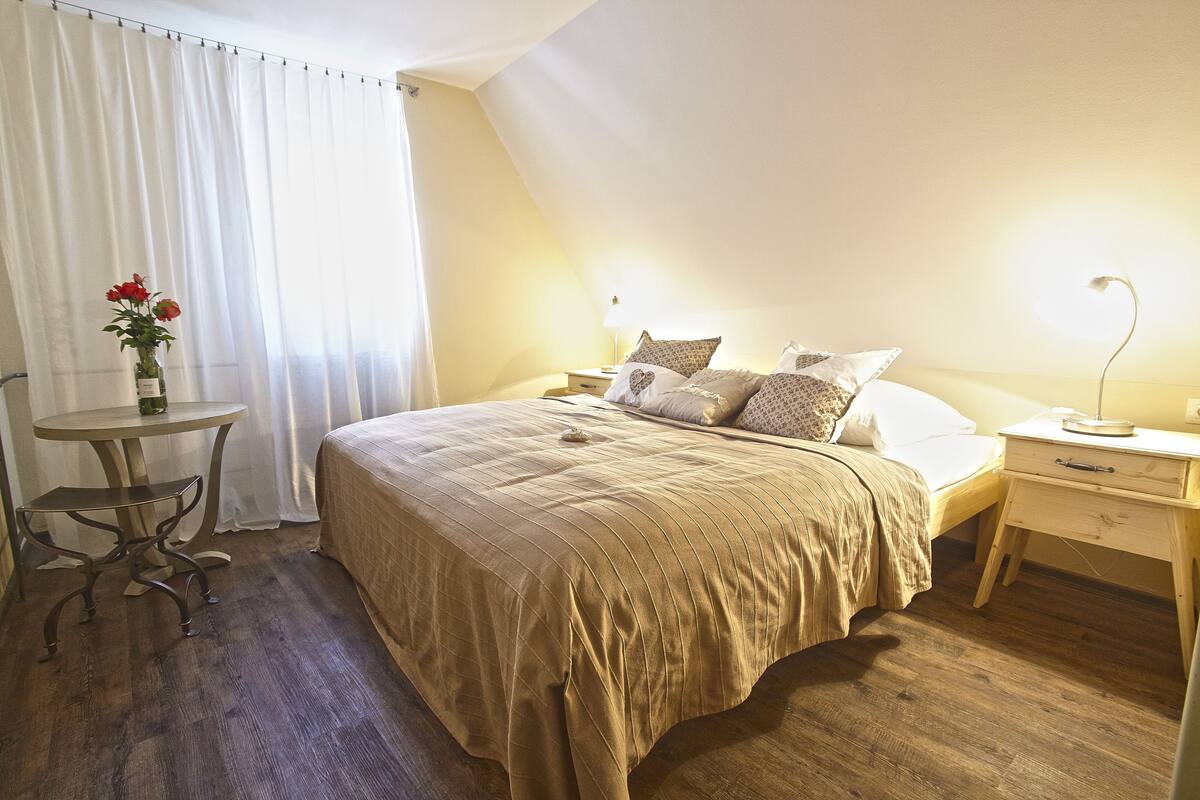Quarto com cama, mesas de cabeceira, uma mesinha com um vaso de flores e uma janela com cortinas.