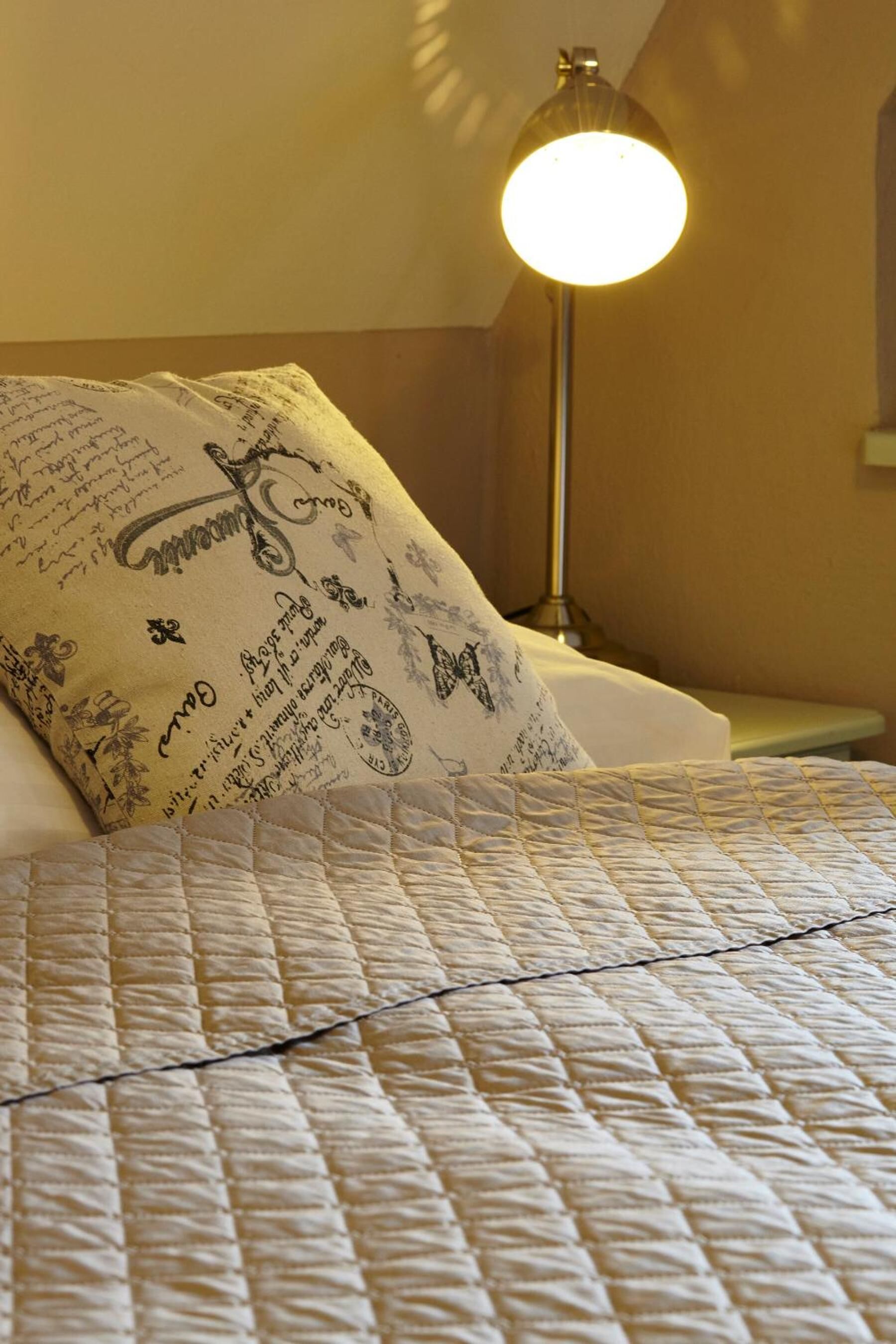 Ein Bett mit gesteppter Decke und einem Kopfkissen mit dekorativem Text. Im Hintergrund ist eine Stehlampe mit runder Schirm zu sehen.