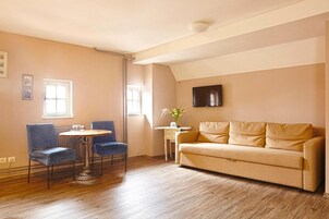 Un salón con un sofá beige, dos sillas azules, una pequeña mesa redonda, una mesita de noche con un florero y un televisor de pantalla plana montado en la pared.