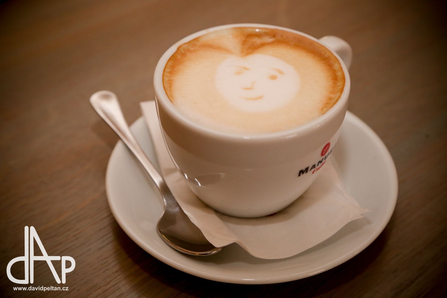 Una taza de café con arte de latte sobre un platito y cuchara, sobre una mesa de madera.