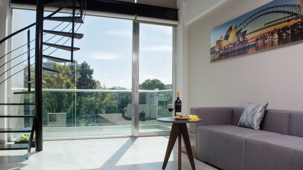 Un soggiorno moderno con un divano grigio, un tavolino in vetro con una bottiglia e degli snack, e un'ampia finestra con vista sulla città e sul verde.