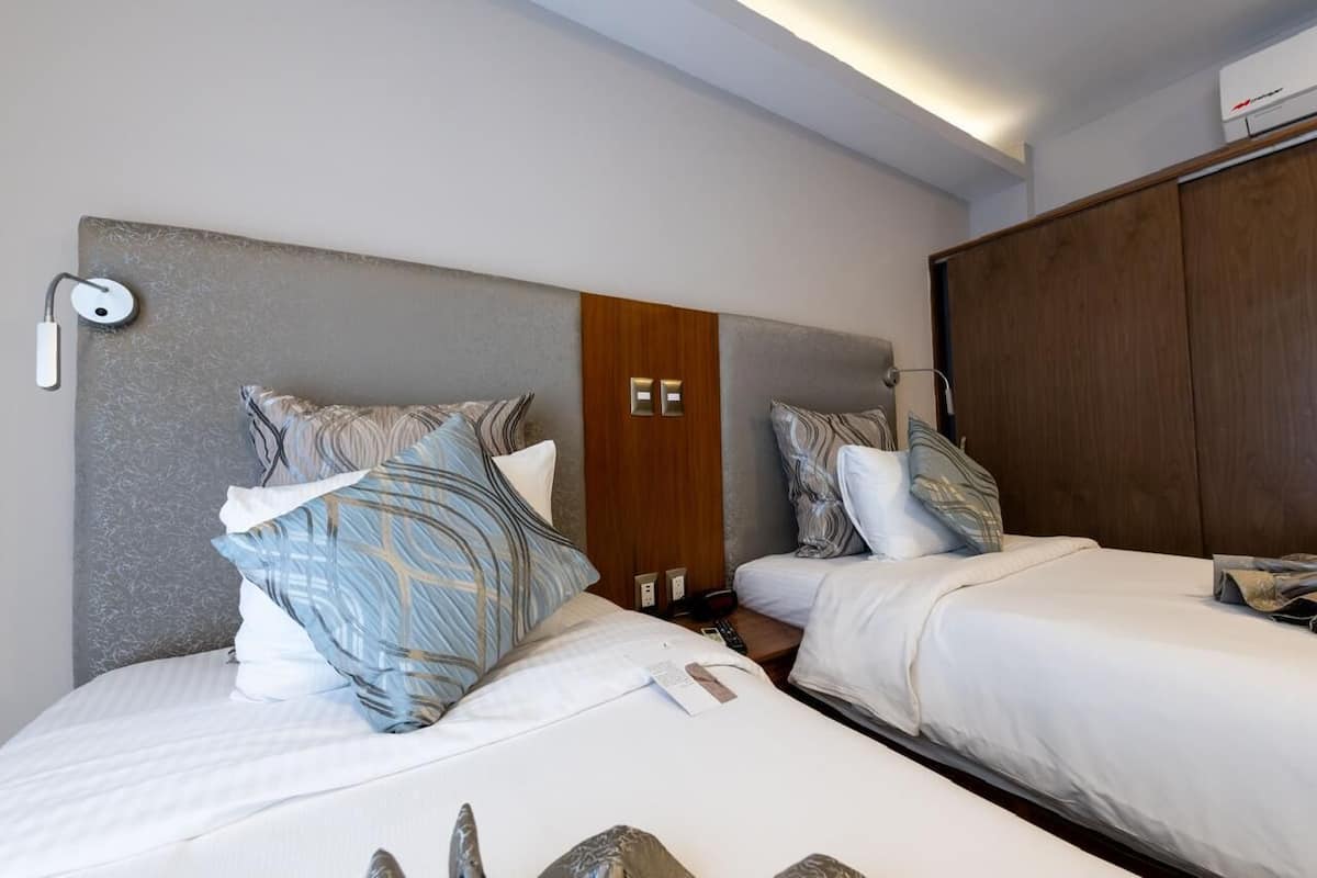 A hotel room with two beds, a wooden wardrobe, a wall-mounted light, and a small nightstand.