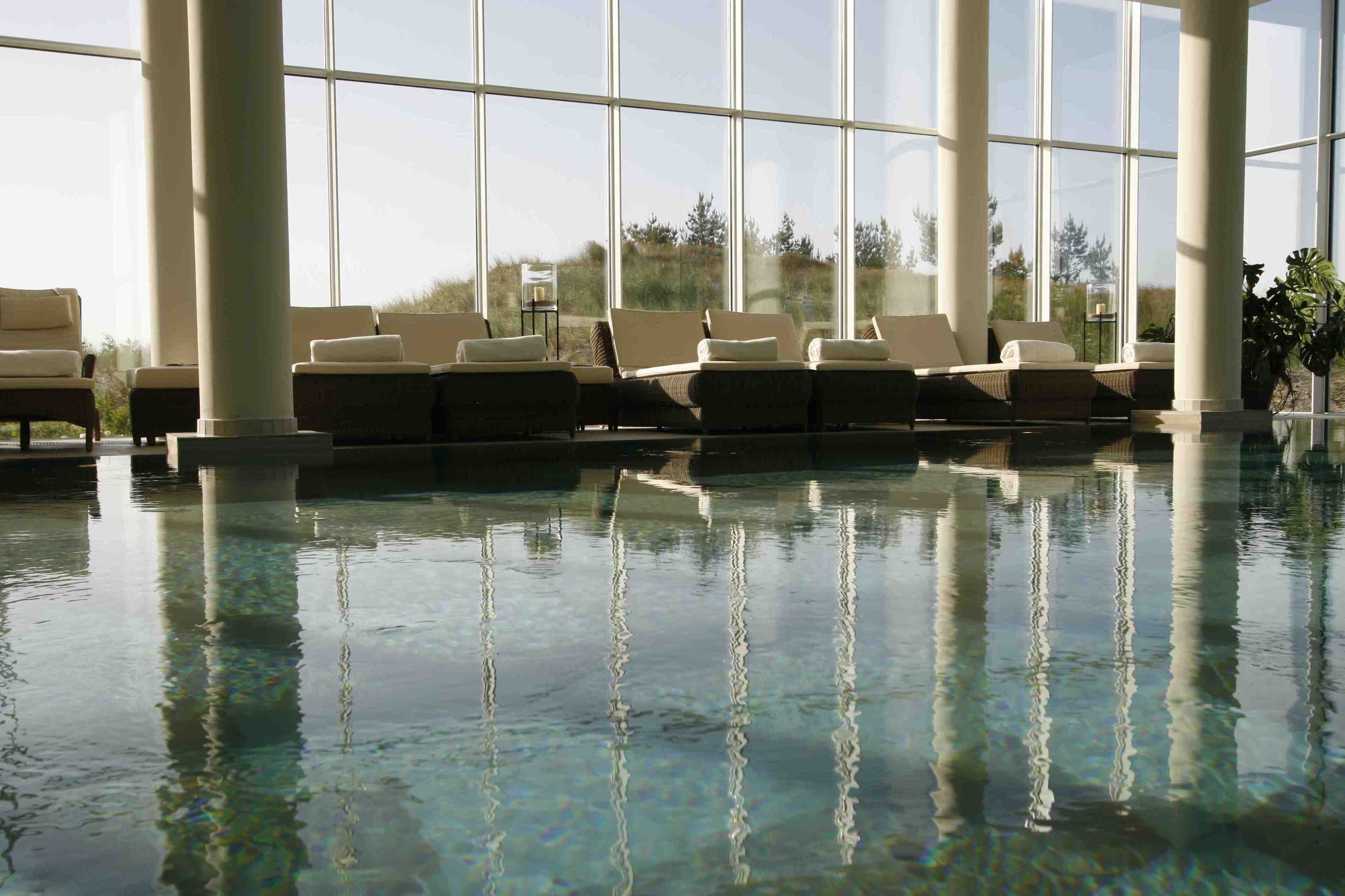 An indoor swimming pool with clear water, large windows, and lounge chairs.