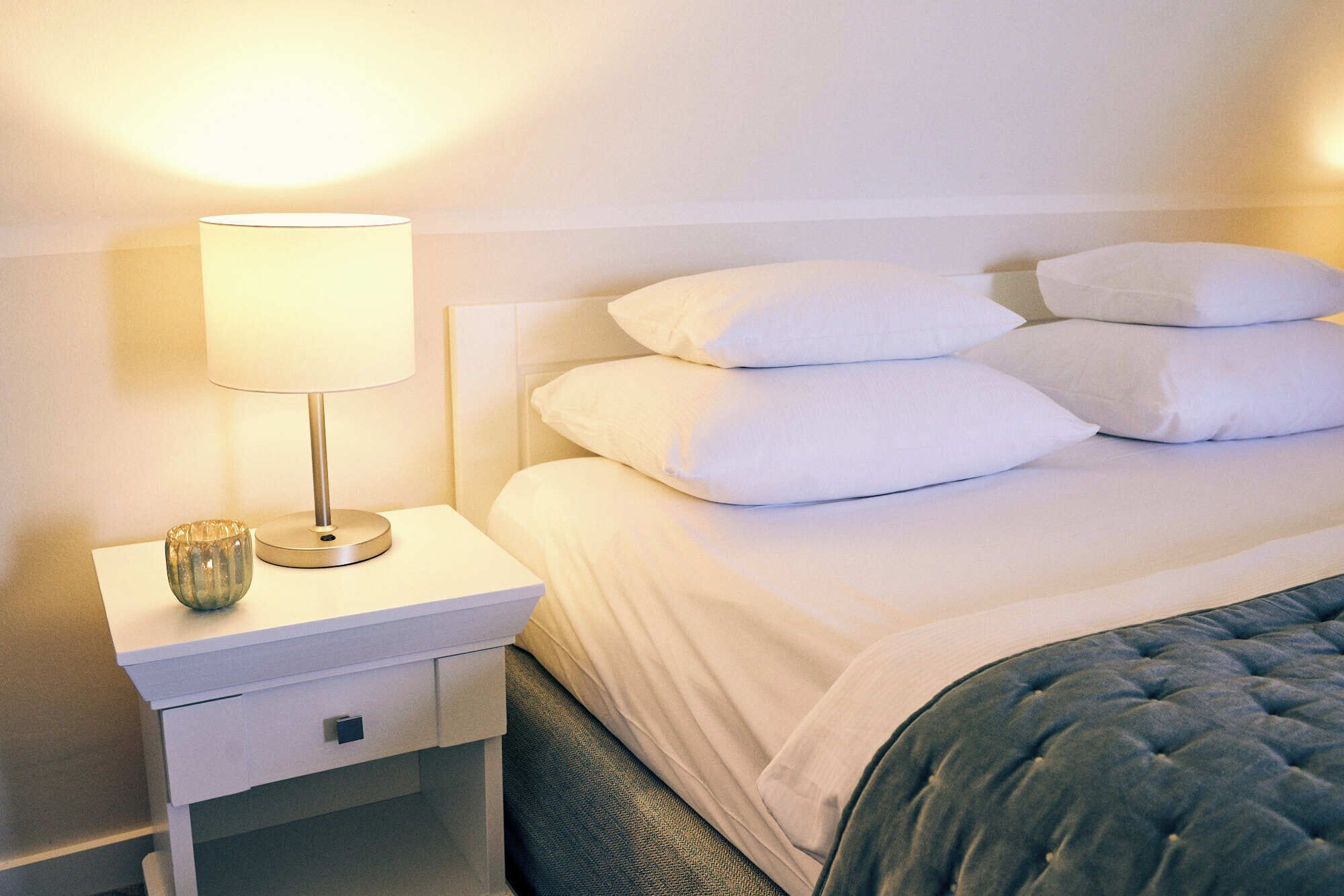 Una cama bien hecha con sábanas blancas y un cabecero tapizado en azul. Junto a la cama, una mesita de noche blanca con una lámpara y un pequeño adorno.