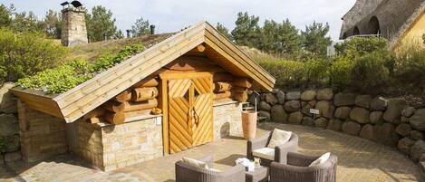 Una cabaña de troncos con chimenea de piedra, puerta de madera y un patio con mobiliario de mimbre.