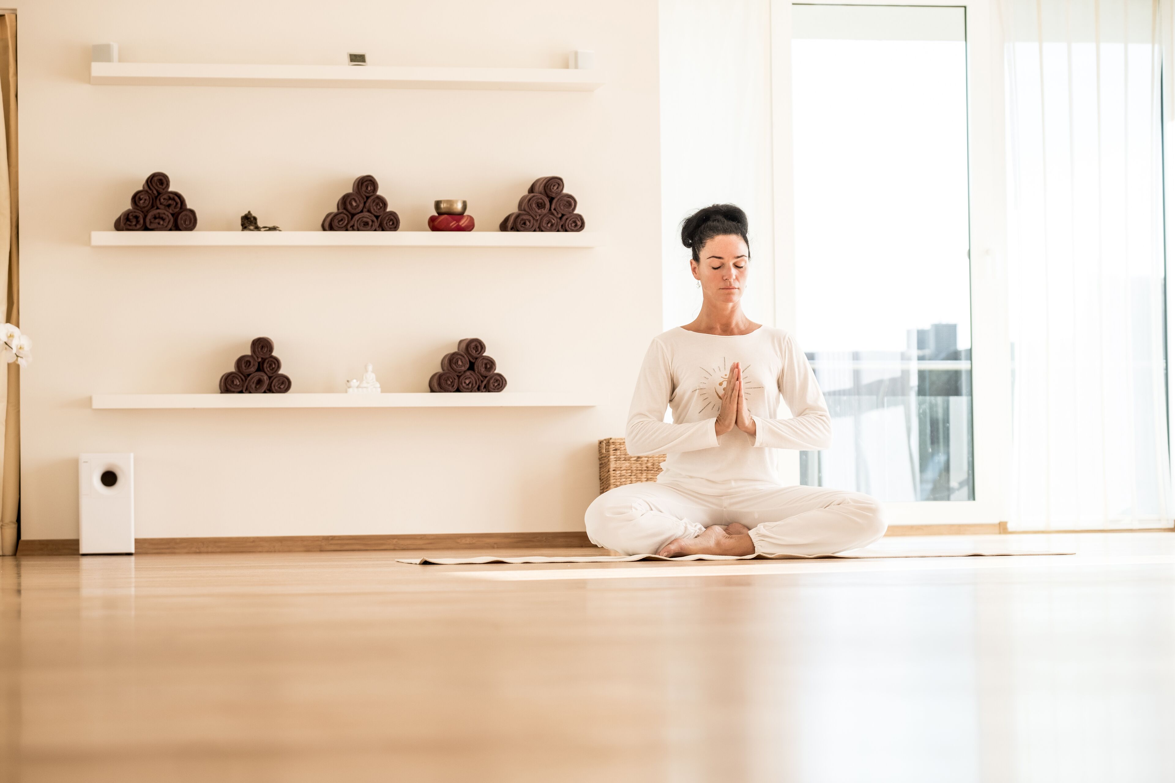 Eine Person meditiert auf einer Yogamatte in einem Raum mit Holzböden, weißen Wänden und Regalen mit aufgerollten Handtüchern und einem Lautsprecher.