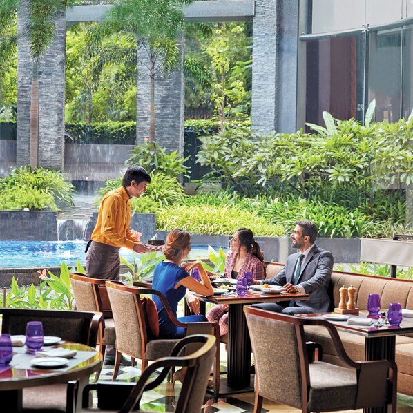 Un restaurante con terraza al aire libre y vistas a una fuente de agua y abundante vegetación.