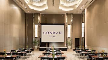 A conference room with a large screen displaying "CONRAD PUNE", equipped with tables, chairs, and a patterned carpet.