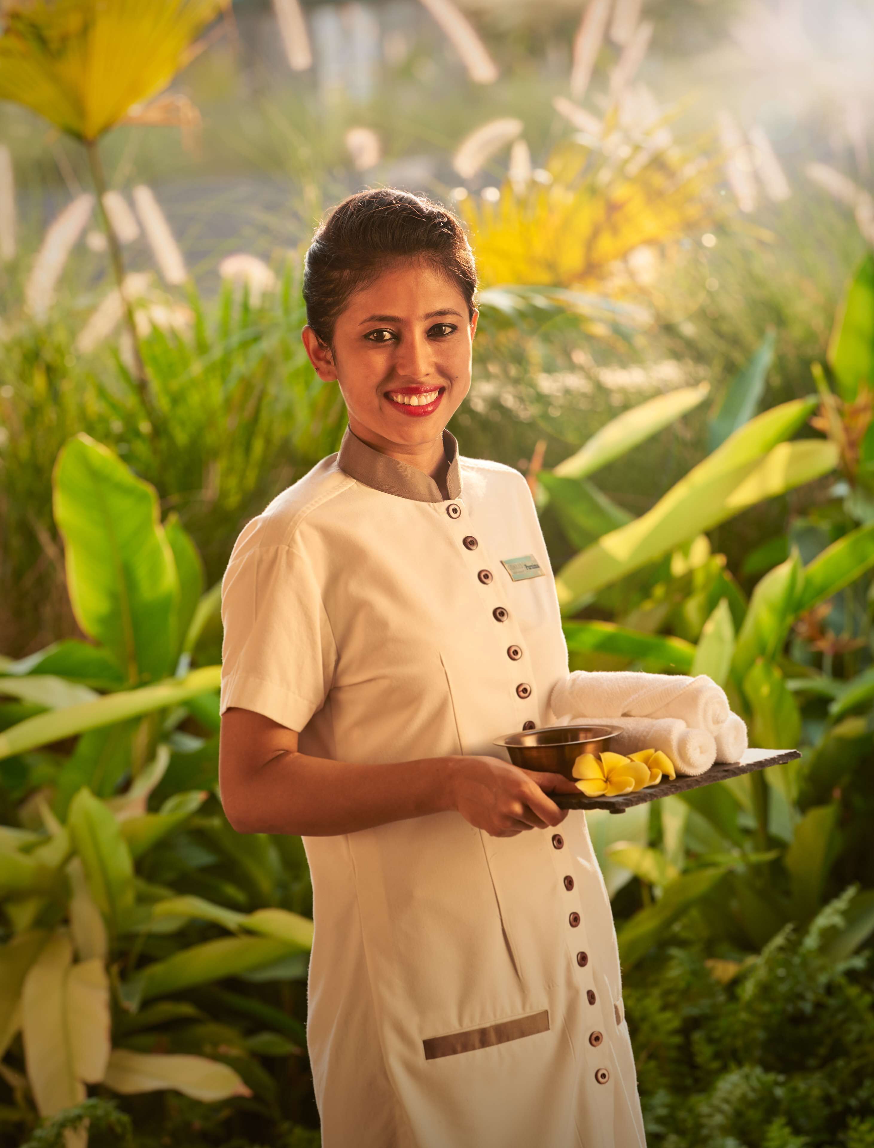 A person in a uniform holding a tray with a bowl and towels, standing in a lush, green outdoor setting.