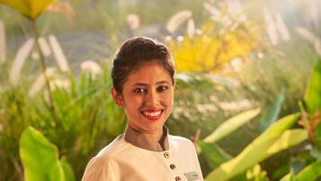 A person in a uniform holding a tray with a bowl and towels, standing in a lush, green outdoor setting.