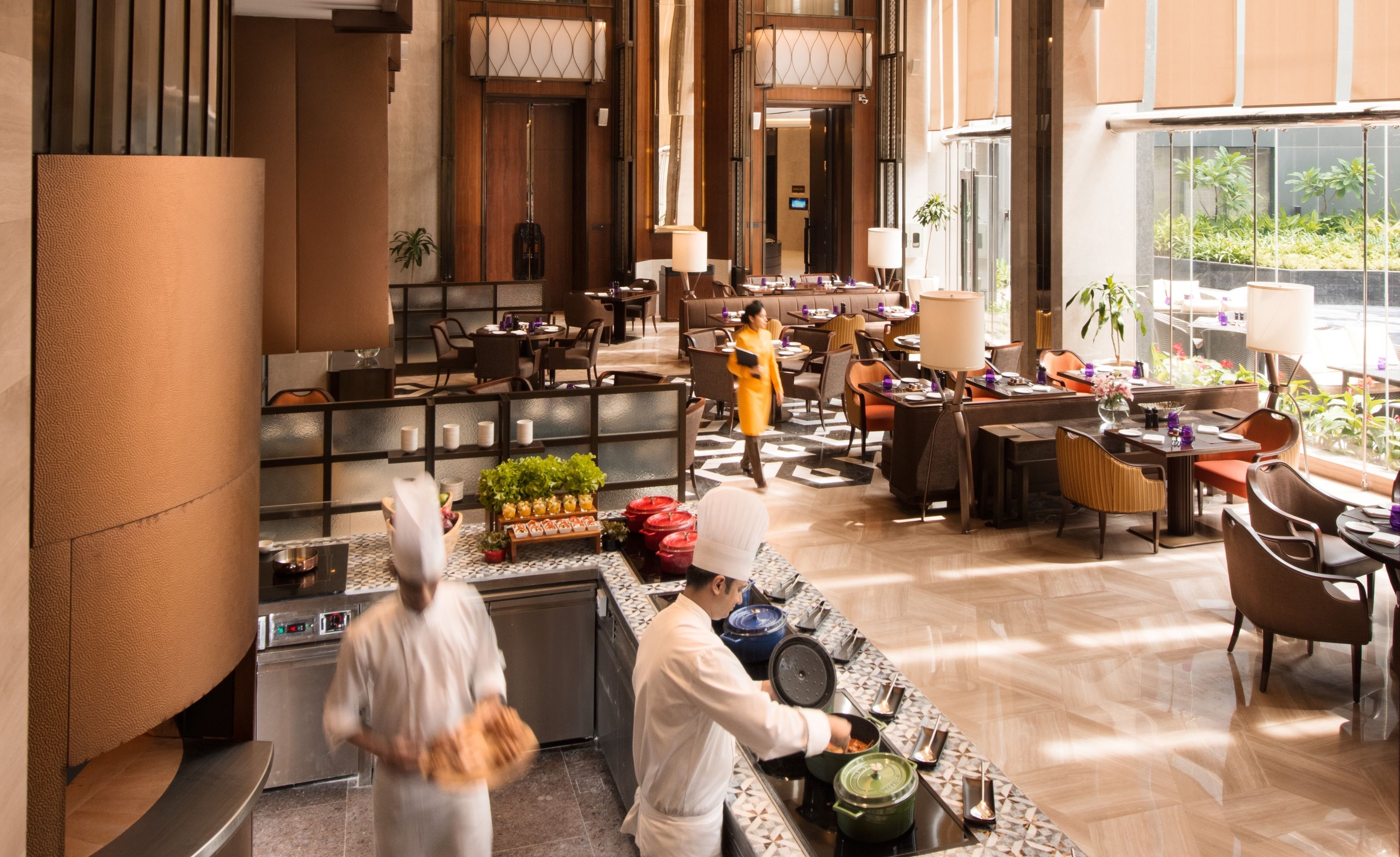 A spacious hotel lobby with multiple dining areas, a central kitchen, and a view of the outdoors.