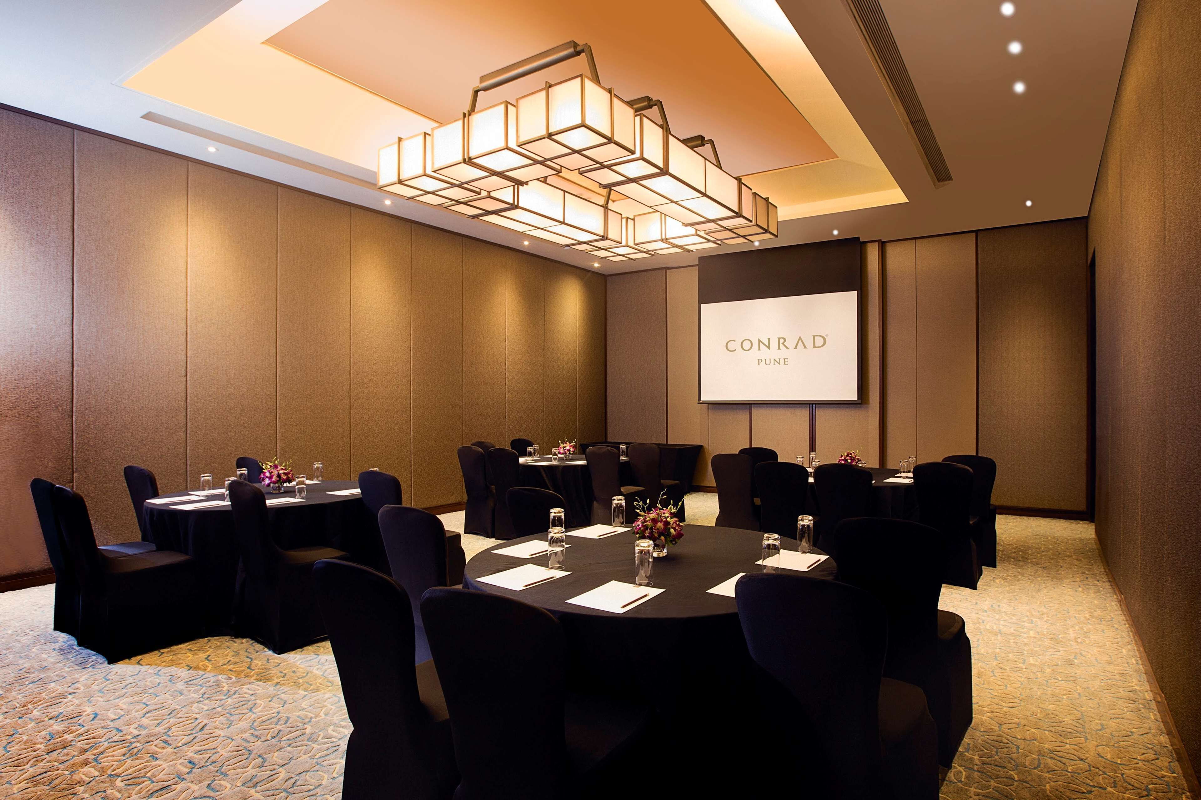Une salle de conférence dotée de plusieurs tables et chaises, d’un écran de projection affichant le texte « CONRAD PUNE » et d’un luminaire moderne au plafond.