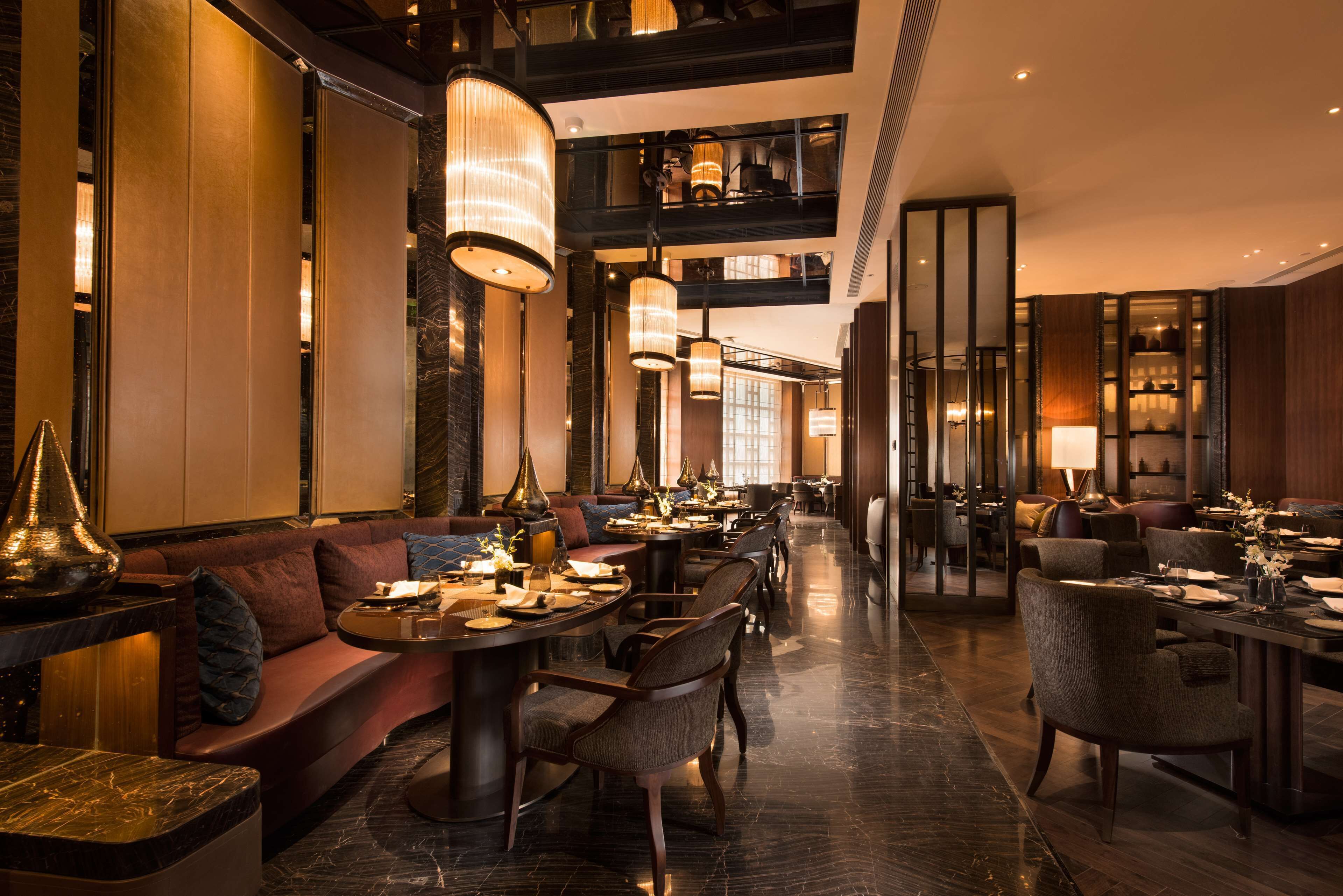 A modern hotel lobby with a central seating area, multiple dining tables, and a high ceiling with hanging lights.