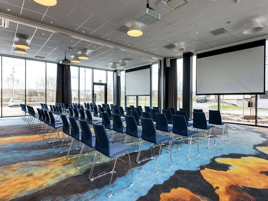 Een vergaderzaal met rijen blauwe stoelen, een groot scherm en een vloerkleed met een kleurrijk abstract patroon.