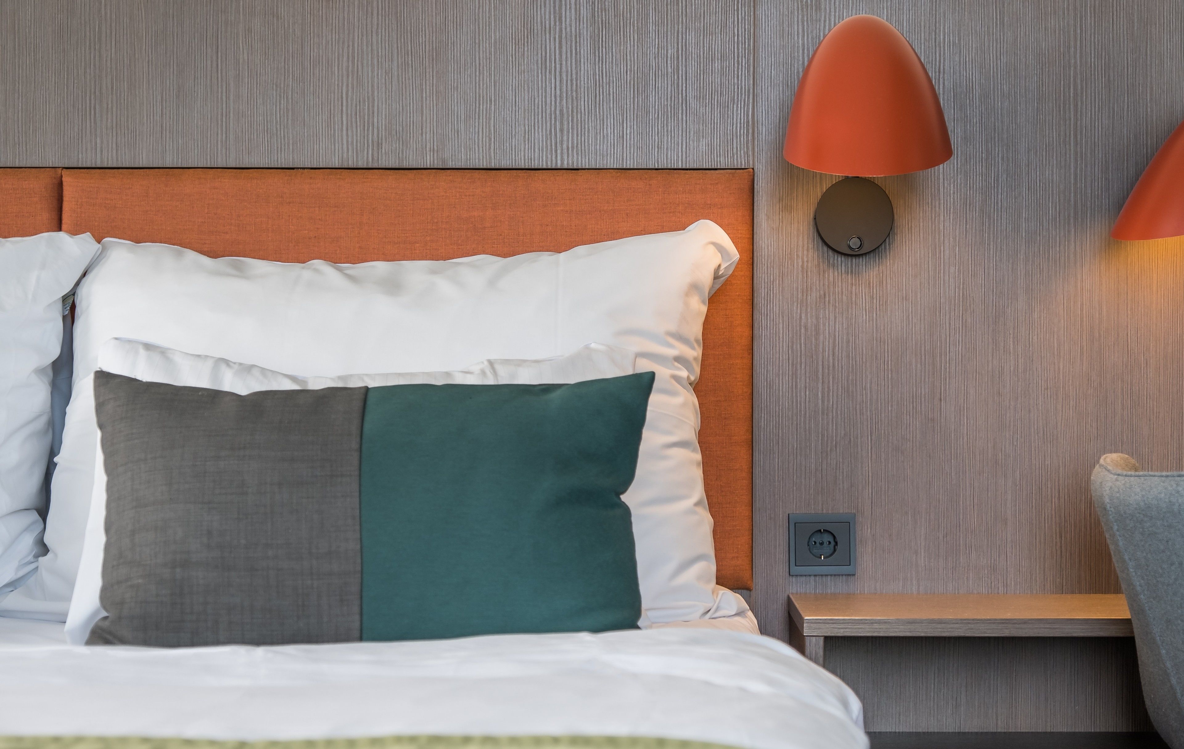 Una cama bien hecha con varios almohadones, un cabecero de madera, una mesita de noche con lámpara y un enchufe.