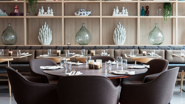 A restaurant with round tables and dark chairs, a wooden wall with shelves of decorative items, and a seating area with cushioned benches. - Quality Hotel Pond (Sandnes)