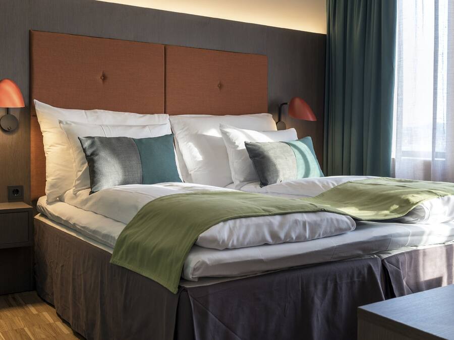 A neatly made bed with a headboard, two bedside lamps, and a view of a window with curtains.