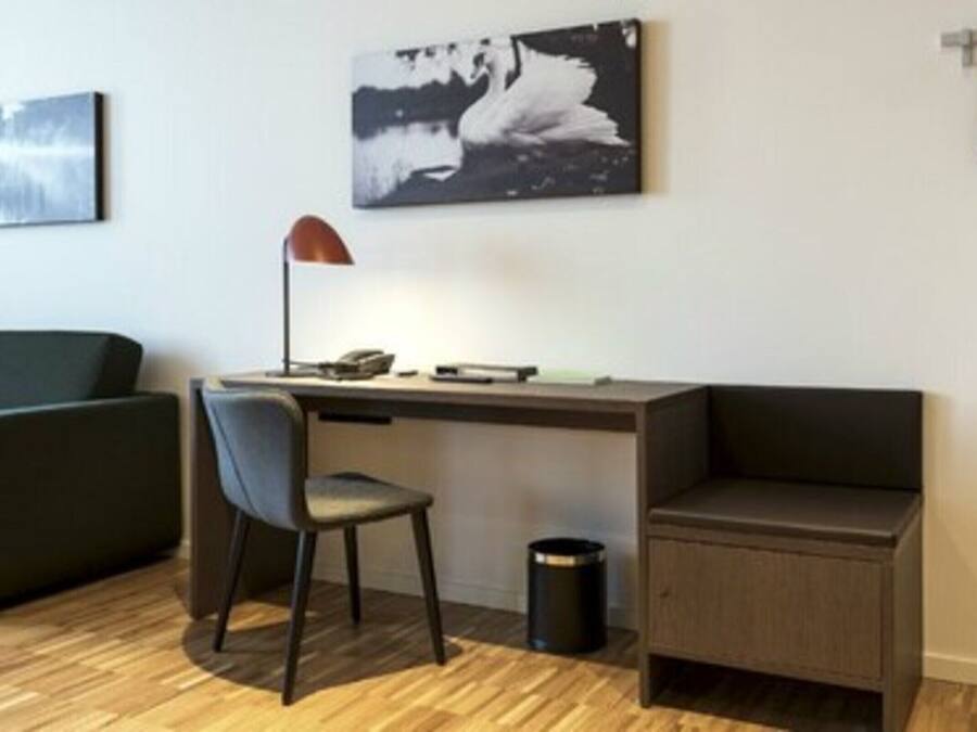 A modern room with a desk, chair, and a small cabinet. There are two wall-mounted artworks and a trash bin.