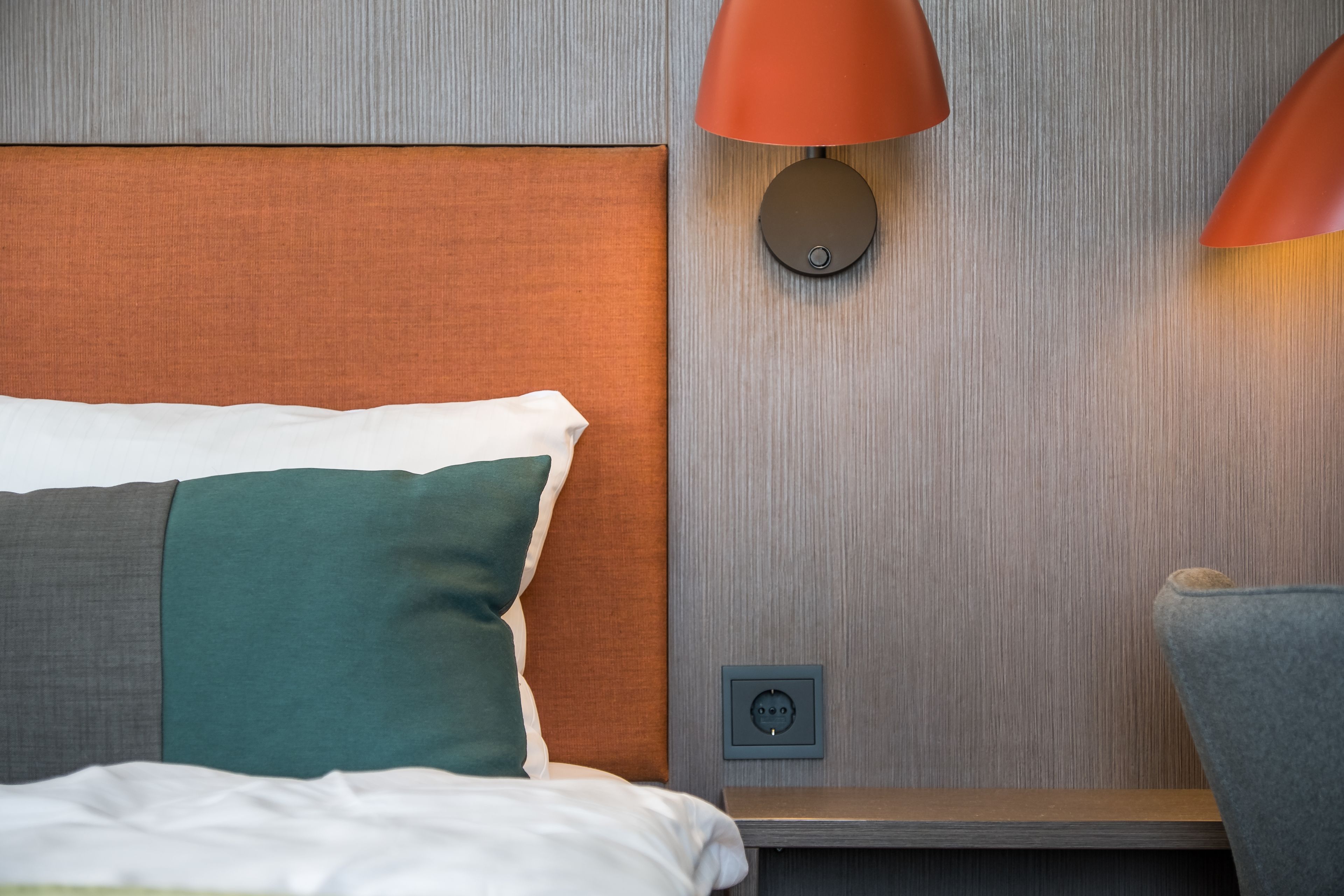A neatly made bed with a headboard, a bedside table with a lamp, and a power outlet on the wall.