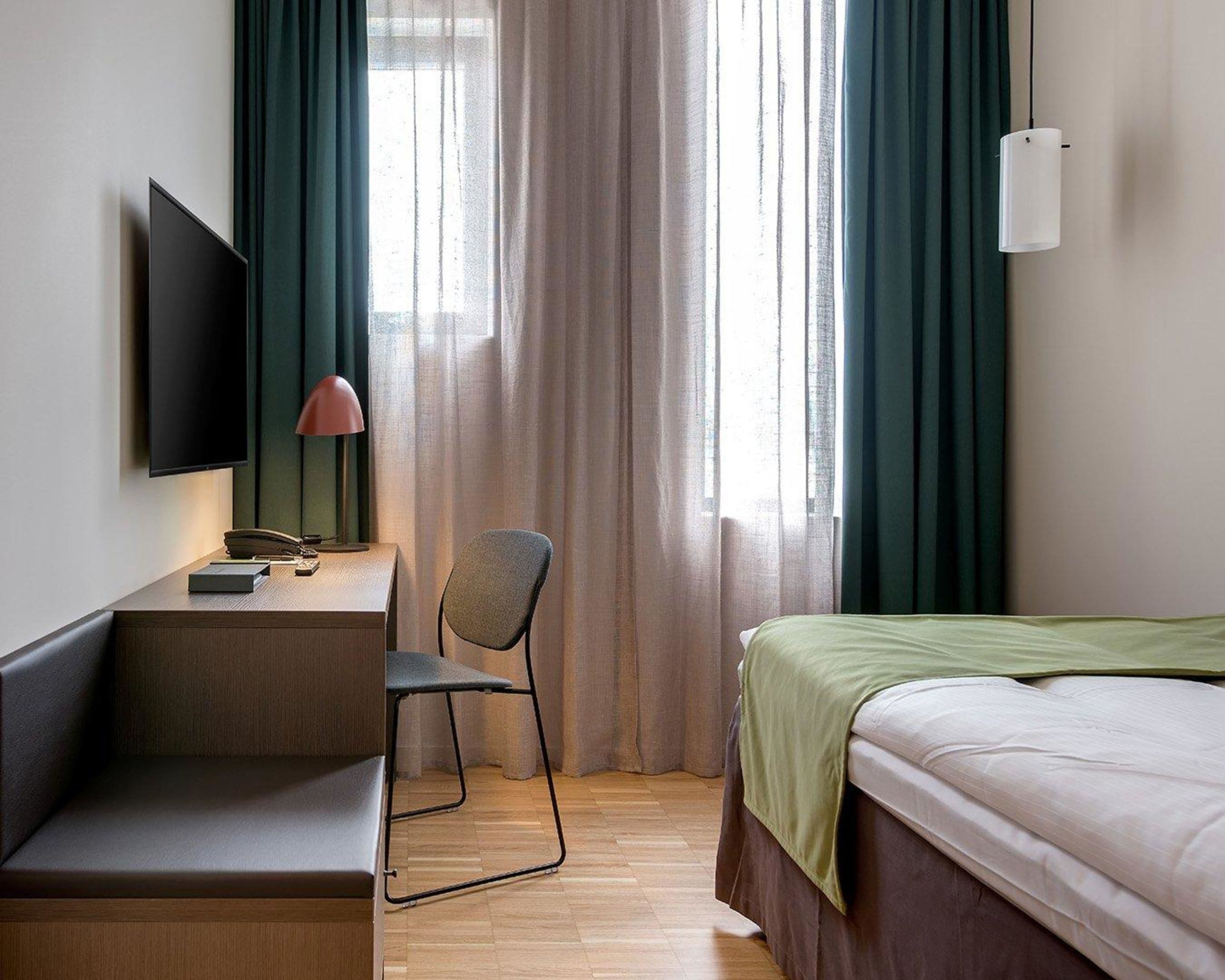 Habitació d'hotel amb un llit, un escriptori amb una cadira, una televisió i una finestra amb cortines.