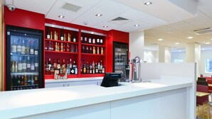 Una zona de bar con mostrador blanco, estanterías rojas con diversas botellas de licor, un refrigerador con productos y un grifo de cerveza.