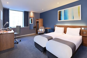Une chambre d’hôtel avec deux lits, un bureau, une chaise, une table ronde et un grand tableau abstrait accroché au mur.