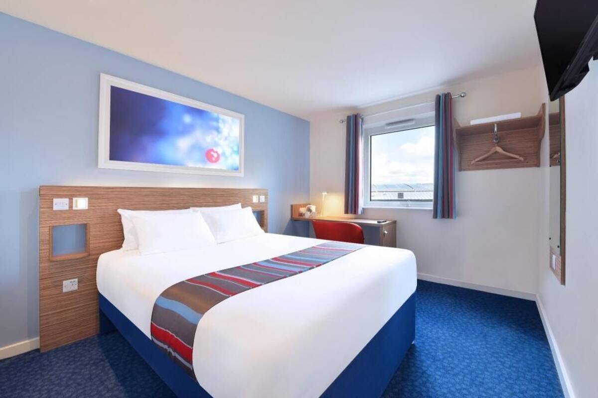 Quarto de hotel com cama, escrivaninha, cadeira, televisão, janela com cortinas e um quadro na parede.
