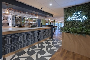 A bar area with a counter, wine glasses, and a neon sign reading "85 bar cafe". - Travelodge Harrogate West Park (Harrogate)