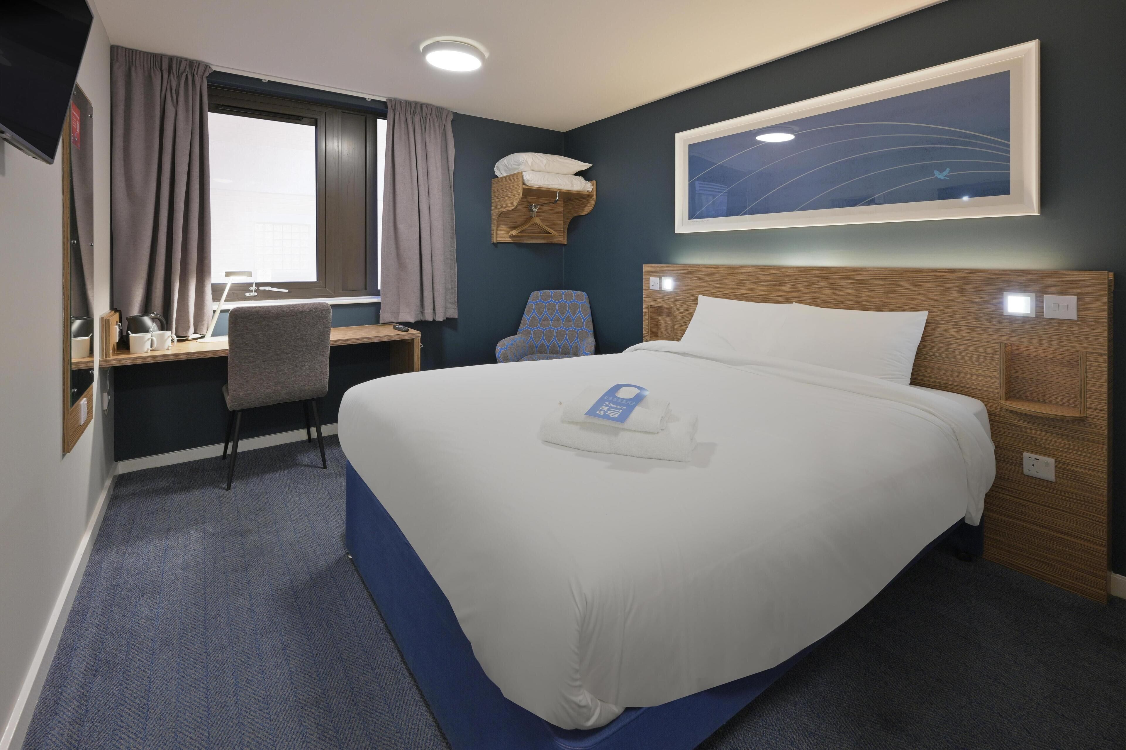 A hotel room with a large bed, a desk with a chair, a window with curtains, a wall-mounted shelf with towels, and a framed picture on the wall.