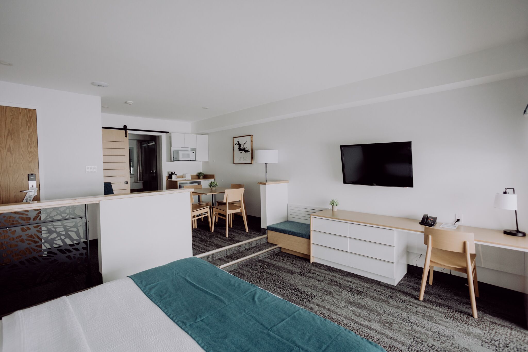A modern hotel room with a bed, desk, and kitchenette.