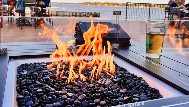 A fire feature with black stones and a caution sign, surrounded by outdoor seating and a view of the water.