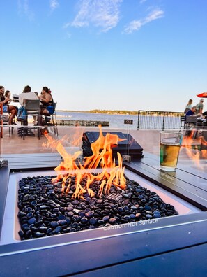 A fire feature with black stones and a caution sign, surrounded by outdoor seating and a view of the water. - La Place Rendez-Vous (Fort Frances)