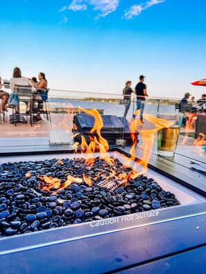 A fire feature with cautionary signage, surrounded by seating and a view of the water. - La Place Rendez-Vous (Fort Frances)
