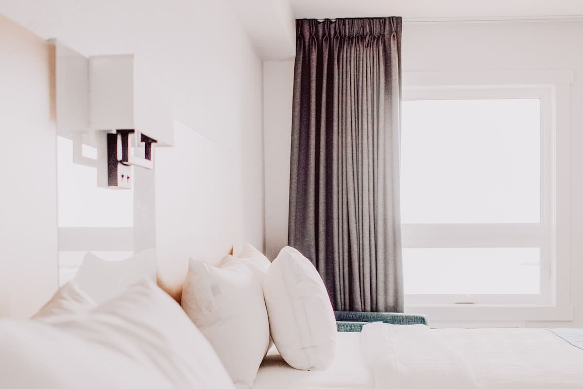 Quarto de hotel com uma cama grande, uma mesinha de cabeceira, janela com cortinas e um espelho na parede.