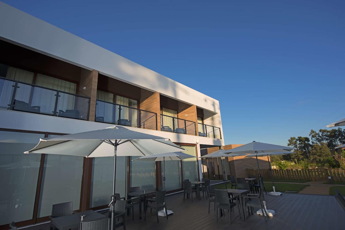 Een modern terras buiten met meerdere tafels en parasols, een houten terras en een gebouw met twee verdiepingen, grote ramen en balkons.