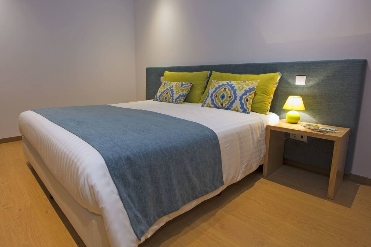 Een netjes opgemaakt bed met een blauwe deken, twee kussens met een geometrisch patroon, en een bedlamp op een houten nachtkastje.
