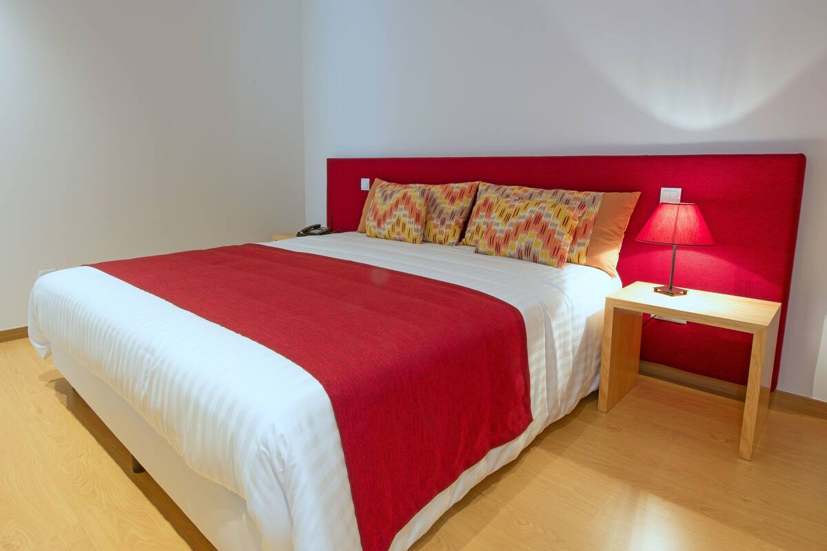 Een bed met een rood hoofdeinde, wit beddengoed en twee gekleurde kussens. Er staat een nachtkastje met een lamp.