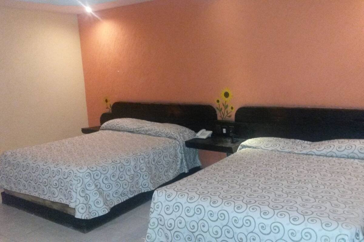 A hotel room with two beds, each with a patterned bedspread, a nightstand with a phone, and a vase with flowers on the wall.