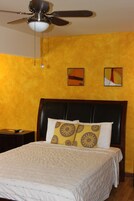 Habitación de hotel con cama, mesita de noche, ventilador de techo y dos obras de arte en las paredes.